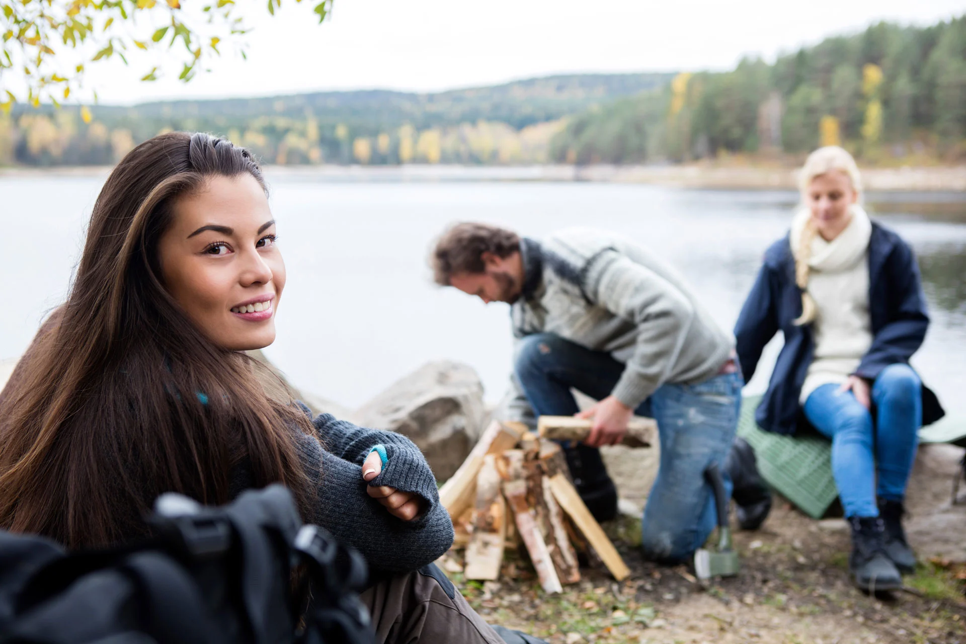 woman smiling at camera with friends preparing a campfire in a serene lakeside setting, possibly a therapeutic outdoor activity for fentanyl addiction recovery