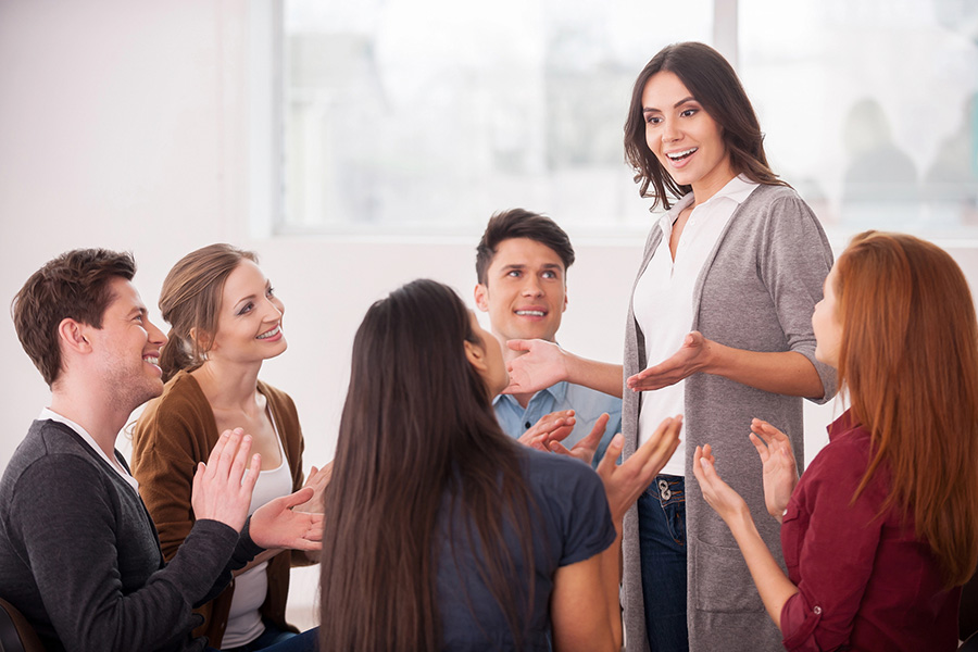 a group claps for a woman standing during a drug or alcohol addiction treatment support meeting.