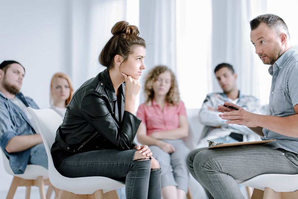a young woman attentively listens during a therapy session with a group of peers and a counselor.