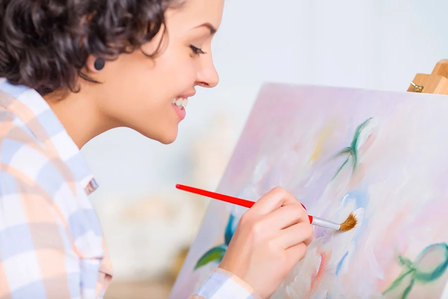 smiling young woman with short curly hair painting on a canvas easel with a paintbrush.
