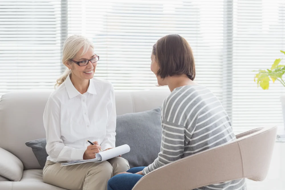 A Smiling Therapist Takes Notes During A Session With A Client, Demonstrating The Professional Care Covered By Bcbsmt. a smiling therapist takes notes during a session with a client, demonstrating the professional care covered by bcbsmt.